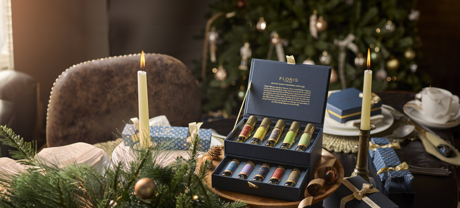 A box of assorted perfumes from The Perfumer's Collection by Floris London, with festive decor in the background, featuring a lit candle and wrapped gifts, makes for the perfect gift for any fragrance connoisseur.