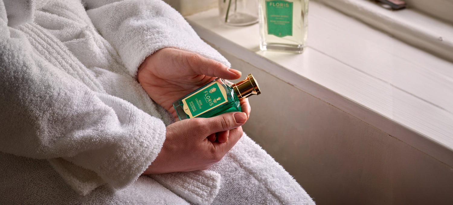 An elegant woman in a bathrobe holding a bottle of Floris Rose Geranium bath essence.