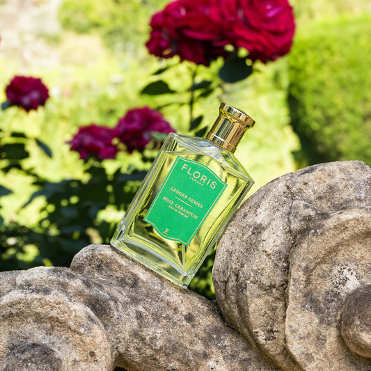 A bottle of Floris London EU Rose Geranium Eau de Parfum sits on a stone sculpture outdoors, with blurred roses and lush greenery in the background.