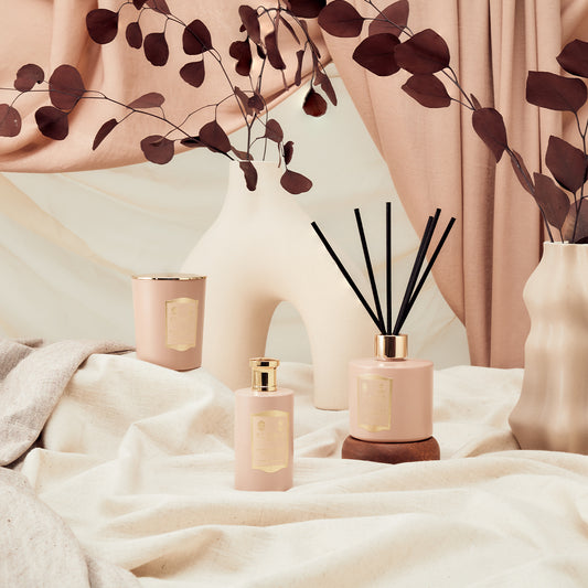 Three blush pink Floris London products in woody Sandalwood & Patchouli scent—a candle, spray, and reed diffuser—sit on beige fabric.