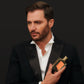 A man in a black suit holds Floris London Santal Intense Eau de Parfum, a sandalwood scent in a rectangular bottle with an orange label, against a dark background.