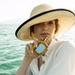 A woman in a wide-brimmed straw hat holds Floris London's Sirena Eau de Parfum by the water, enjoying its fresh floral scent on a sunny day.