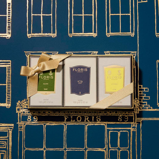 The Soap Collection | For Him by Floris London EU: three signature soaps with a gold ribbon, set against a blue and gold storefront illustration.