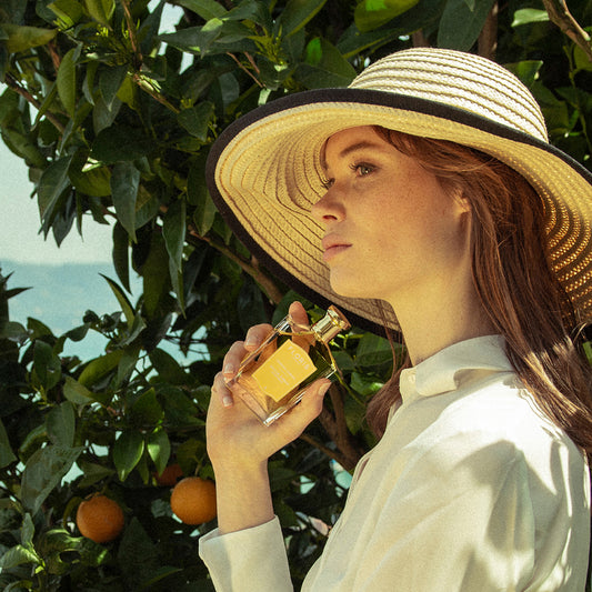 A woman in a wide-brimmed hat holds Floris London EU’s Soulle Ámbar perfume near her face, standing by an orange tree with fruit behind her.