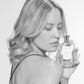 A woman with light wavy hair holds a Floris London EU Stephanotis perfume bottle close to her face, enjoying its elegant floral scent against a plain background.