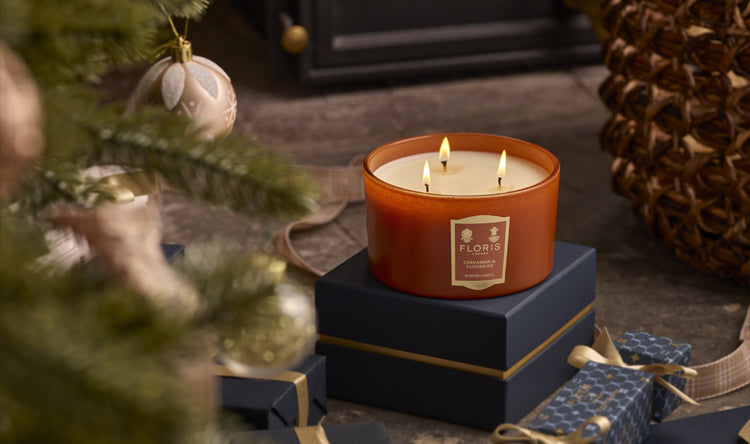 A limited-edition Cinnamon & Tangerine Deluxe Three Wick Candle by Floris London sits atop a stack of gifts near a decorated Christmas tree, with a fireplace glowing warmly in the background.