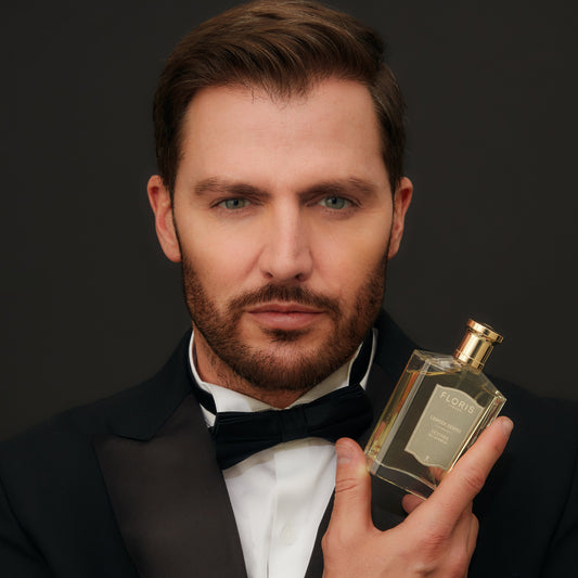 A man in a tuxedo holds a bottle of Floris London EU Vetiver fragrance close to his face against a plain dark background.