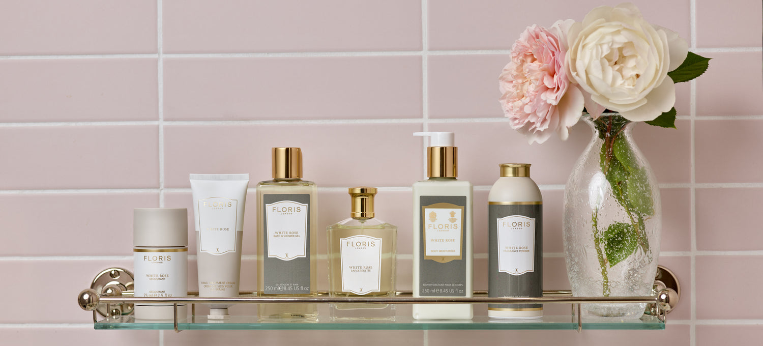 The White Rose collection displayed on a glass shelf with a vase of roses in a pink tiled bathroom.