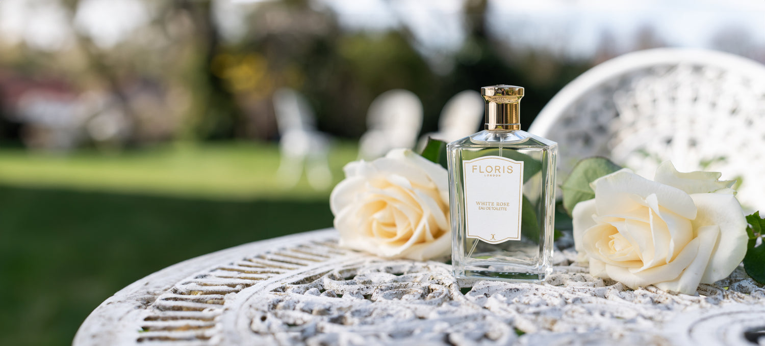 A bottle of White Rose Eau de Toilette on a white iron table, surrounded by white roses in a garden.