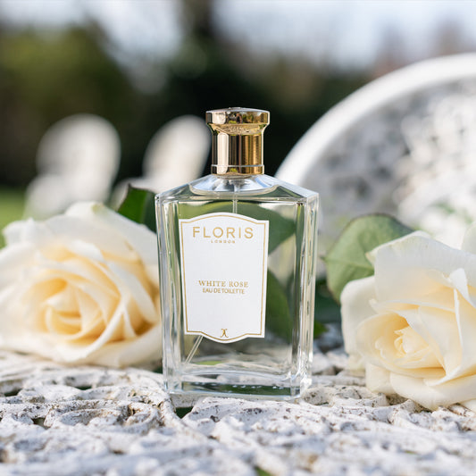 A clear glass bottle of Floris London White Rose - Eau de Toilette rests on a white surface beside two roses, set against a blurred outdoor background.